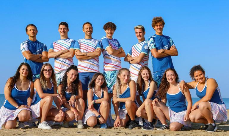 Lächelnde Gruppe am Strand, blauer Himmel und fröhliche Sommerstimmung.