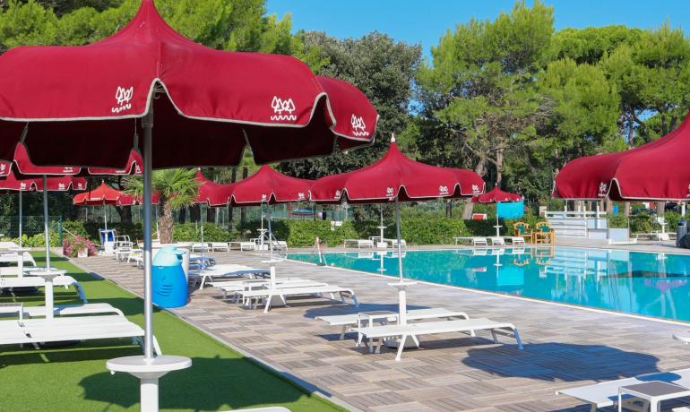 Piscina con lettini e ombrelloni rossi, giornata estiva e cielo limpido.